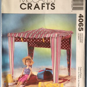 May include: A McCall's Crafts sewing pattern envelope featuring a doll and a miniature beach scene. The scene includes a striped canopy, a doll in pink pants, a beach bag, and other accessories. The text "McCall's Crafts" is at the top.