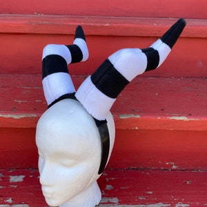 May include: Black and white striped headband with two horns. The horns are made of fabric and are shaped like twisted legs. The headband is on a white mannequin head.