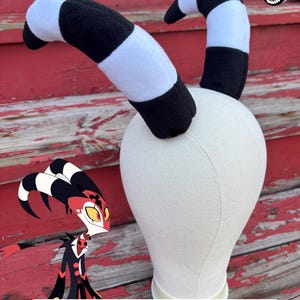 May include: A pair of plush, curved horns featuring alternating black and white horizontal stripes, mounted on a beige mannequin head. The horns are inspired by the character Blitzo, with a digital illustration of the character and the text Blitzo Horns Printable Pattern visible.