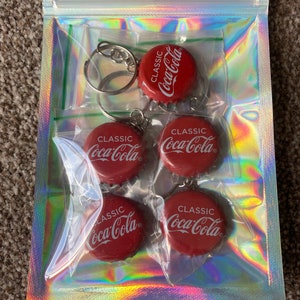 Puede incluir: Una bolsa transparente iridiscente con cremallera que contiene cinco llaveros de tap&oacute;n de botella de Coca-Cola rojos. Cada llavero presenta el logotipo "CLASSIC Coca-Cola" en blanco. Los llaveros est&aacute;n unidos a llaveros plateados.