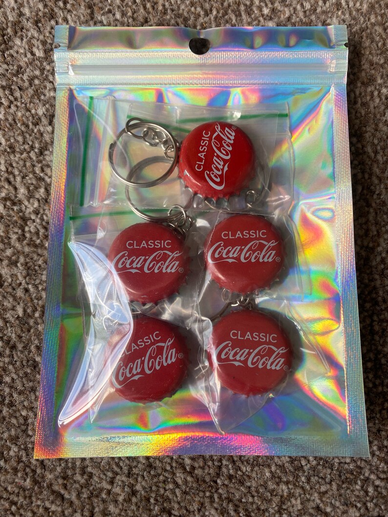 Puede incluir: Una bolsa transparente iridiscente con cremallera que contiene cinco llaveros de tap&oacute;n de botella de Coca-Cola rojos. Cada llavero presenta el logotipo "CLASSIC Coca-Cola" en blanco. Los llaveros est&aacute;n unidos a llaveros plateados.