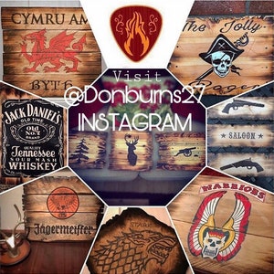 May include: A collection of rustic wooden signs with various designs. The signs feature images of a dragon, a skull, a deer, a cannon, and text including "CYMRU AM", "The Jolly Roger", "Jack Daniel's", "Saloon", "J&auml;germeister", "STARK", and "WARRIORS".