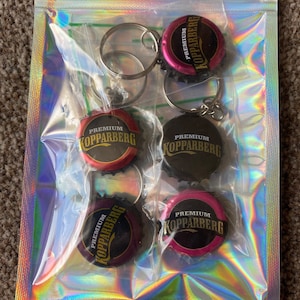 Puede incluir: Una bolsa de pl&aacute;stico transparente contiene cinco llaveros. Cada llavero presenta una chapa de botella con el texto "Premium Kopparberg" en letras doradas. Las chapas son de varios colores, incluyendo rojo, negro y rosa, y est&aacute;n unidas a llaveros plateados.