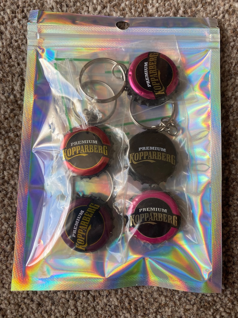 Puede incluir: Una bolsa de pl&aacute;stico transparente contiene cinco llaveros. Cada llavero presenta una chapa de botella con el texto "Premium Kopparberg" en letras doradas. Las chapas son de varios colores, incluyendo rojo, negro y rosa, y est&aacute;n unidas a llaveros plateados.