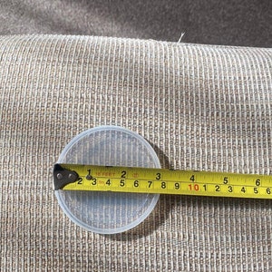 May include: A clear, round plastic container is measured with a tape measure. The container is approximately 4 inches in diameter. The tape measure shows measurements in inches and feet.