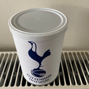 May include: A white cylindrical tumbler with a gray lid, featuring the Tottenham Hotspur logo in navy blue. The logo includes a stylized rooster atop a soccer ball, with the team name printed below. The tumbler is likely made of plastic.