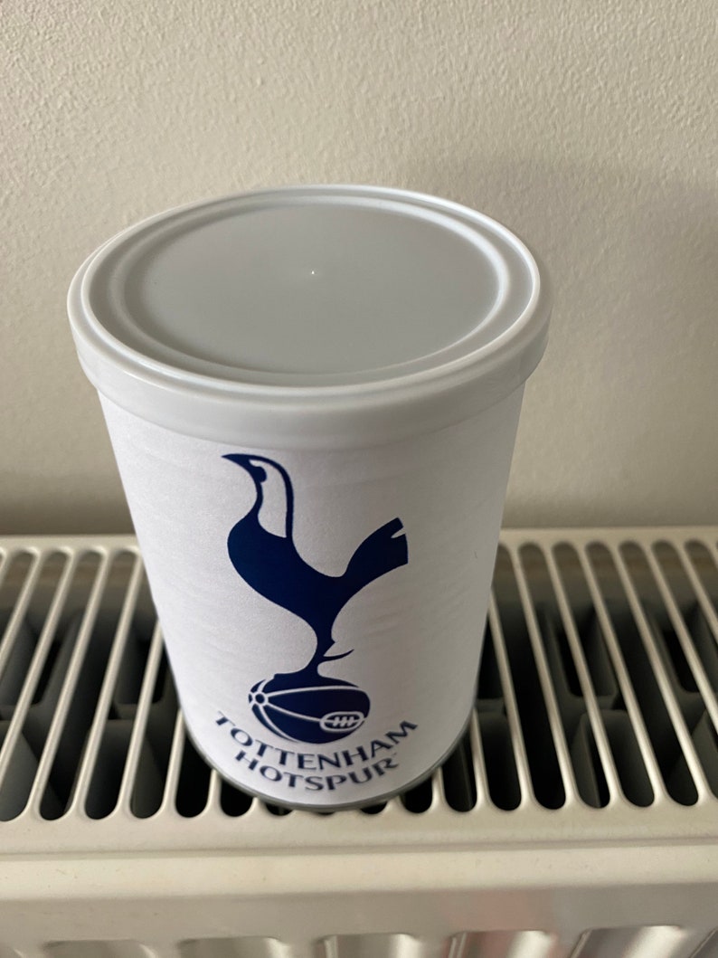 May include: A white cylindrical tumbler with a gray lid, featuring the Tottenham Hotspur logo in navy blue. The logo includes a stylized rooster atop a soccer ball, with the team name printed below. The tumbler is likely made of plastic.