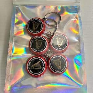 Puede incluir: Llaveros con dijes de tapones de botella. Los tapones son rojos y negros, con el texto "THE ORIGINAL EXTRA STOUT" y un arpa dorada. Los llaveros son plateados y los dijes est&aacute;n empaquetados en una bolsa transparente iridiscente.