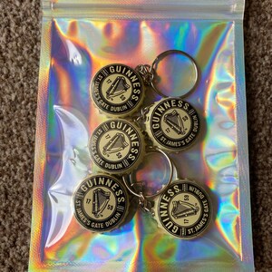 Puede incluir: Un conjunto de cinco llaveros de tap&oacute;n de botella Guinness en una bolsa con cremallera hologr&aacute;fica. Cada llavero presenta el logotipo de Guinness con un arpa y el texto "St. James's Gate Dublin".