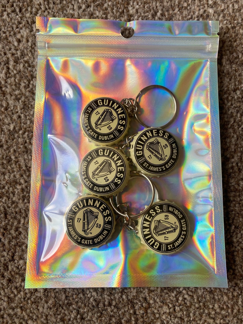 Puede incluir: Un conjunto de cinco llaveros de tap&oacute;n de botella Guinness en una bolsa con cremallera hologr&aacute;fica. Cada llavero presenta el logotipo de Guinness con un arpa y el texto "St. James's Gate Dublin".