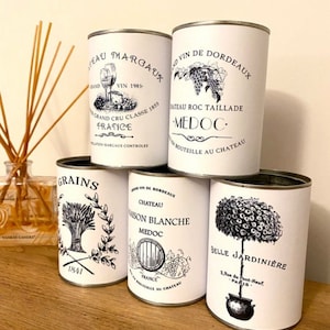 May include: Five white metal cans with black and white vintage labels. The labels feature images of grapes, wheat, a tree, and a wine glass. The text on the labels includes the words "Chateau", "Medoc", "France", and "Grains".