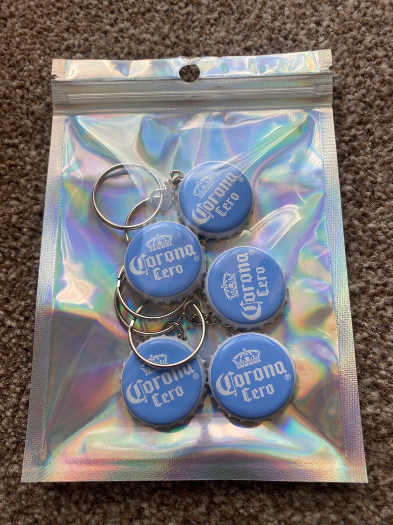Puede incluir: Una bolsa de pl&aacute;stico transparente e iridiscente que contiene cinco llaveros. Cada llavero presenta una tapa de botella azul claro con el logotipo "Corona Cero" en blanco. Los llaveros tienen anillos de metal plateado.