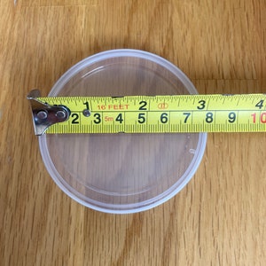 May include: A clear, round plastic container with a white rim is measured with a yellow tape measure. The tape measure shows the container is approximately 3 inches in diameter. The container sits on a wooden surface.