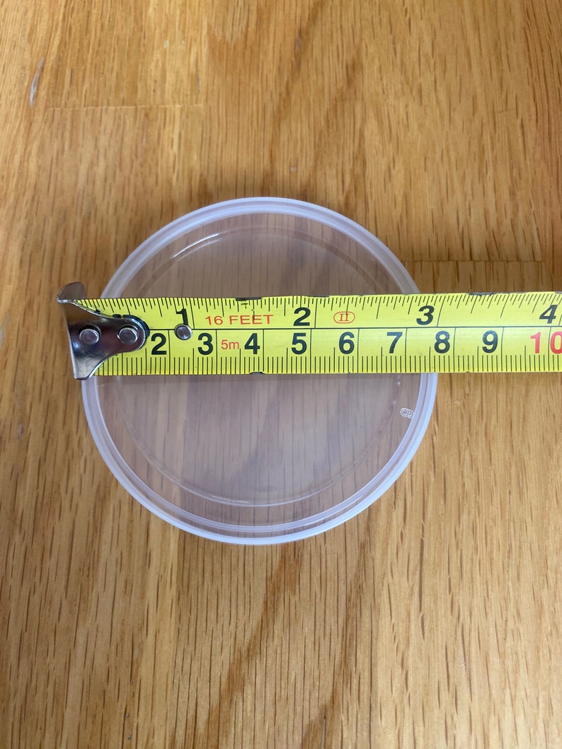 May include: A clear, round plastic container with a white rim is measured with a yellow tape measure. The tape measure shows the container is approximately 3 inches in diameter. The container sits on a wooden surface.