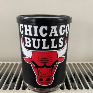 May include: A black glass with the Chicago Bulls logo. The logo features a red bull head with white horns and the words "CHICAGO BULLS" in white block letters. The glass is sitting on a white surface.