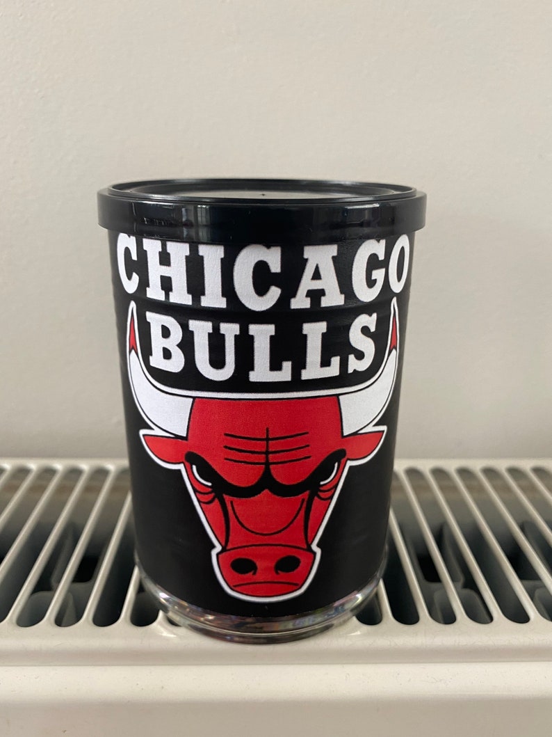 May include: A black glass with the Chicago Bulls logo. The logo features a red bull head with white horns and the words "CHICAGO BULLS" in white block letters. The glass is sitting on a white surface.