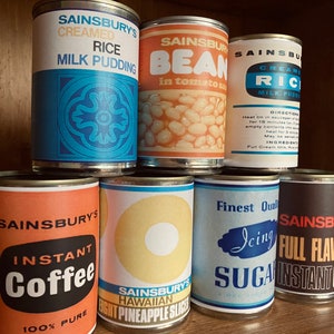 Puede incluir: Una colección de latas de alimentos Sainsbury's vintage, que incluyen arroz con leche, frijoles en salsa de tomate, café instantáneo, rodajas de piña y azúcar glas. Las latas presentan diseños retro con texto en negrita y etiquetas coloridas.