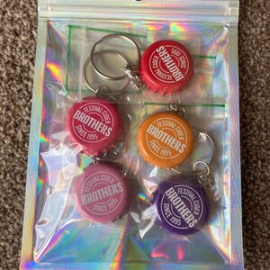Puede incluir: Una bolsa de pl&aacute;stico transparente que contiene cinco llaveros con forma de tap&oacute;n de botella. Cada llavero tiene un tap&oacute;n de diferente color: rojo, rosa, naranja, rosa claro y morado. Los tapones tienen el texto "FESTIVAL CIDER BROTHERS SINCE 1995".