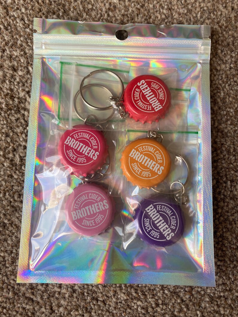 Puede incluir: Una bolsa de pl&aacute;stico transparente que contiene cinco llaveros con forma de tap&oacute;n de botella. Cada llavero tiene un tap&oacute;n de diferente color: rojo, rosa, naranja, rosa claro y morado. Los tapones tienen el texto "FESTIVAL CIDER BROTHERS SINCE 1995".
