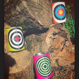 May include: Three tin cans with bullseye targets printed on them. The targets are in different colors: red, yellow, and pink. The cans are sitting on a pile of tree logs.