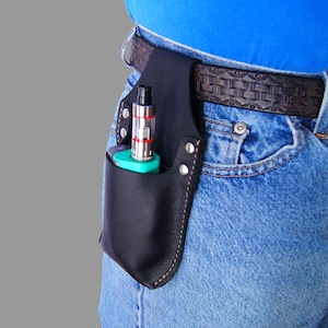 May include: A black leather belt holster with a stitched edge, designed to hold a vaping device. The holster is worn on a person's hip, secured with a brown leather belt. A black and green vaping device is inserted into the holster.