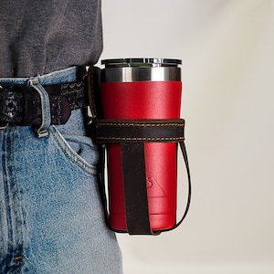 May include: A brown leather belt holster for a red insulated tumbler with a black lid. The holster has a strap that goes around the waist.