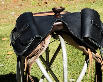 May include: A black leather saddle with two saddlebags on either side. The saddle is sitting on a metal stand.