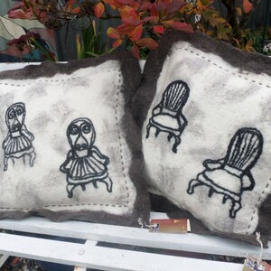 May include: Two white throw pillows with brown trim. One pillow features a black embroidered design of two chairs. The other pillow features a black embroidered design of two figures sitting in chairs.