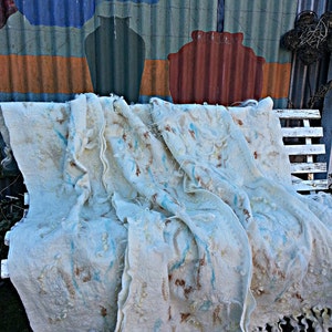 May include: A white, hand-woven throw blanket with a light blue and brown pattern. The blanket has a fringe border and is draped over a white bench.