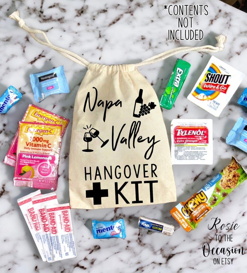 May include: A white canvas bag with a black drawstring, printed with the text "Napa Valley Hangover + Kit". The bag is filled with various items, including a bottle of wine, a glass of wine, a bottle of paracetamol, a pack of chewing gum, a pack of Emergen-C, a pack of plasters, and a granola bar.