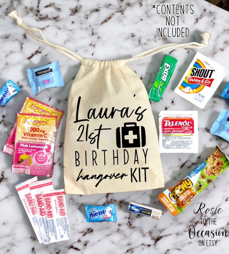 Op de afbeelding: Een witte canvas tas met een zwart trekkoord, bedrukt met de tekst "Laura's 21st Birthday hangover KIT" en een zwart EHBO-doosicoon. De tas is gevuld met verschillende items, waaronder een roze en wit pakje Emergen-C, een wit pakje Tylenol, een groen pakje Extra kauwgom, een wit pakje Shout Wipe & Go, een wit pakje Band-Aid, een blauw pakje Tums, een blauw en wit pakje Mentos, en een bruine en oranje Chewy chocolade-granola reep.