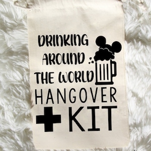 K&ouml;nnte beinhalten: Ein wei&szlig;er Baumwollbeutel mit Kordelzug und schwarzem Text, der "Drinking Around The World Hangover Kit" lautet, mit einem schwarzen Pluszeichen und dem Wort "KIT" in gro&szlig;en Buchstaben. Neben dem Text befindet sich eine schwarze Silhouette eines Bierkruges mit einer Mickey-Maus-Kopf oben drauf.