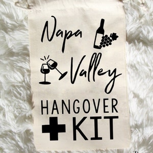 May include: White canvas drawstring bag with black text and graphics that reads "Napa Valley Hangover Kit" with a wine bottle, grapes, and two wine glasses.