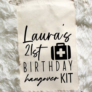 Op de afbeelding: Een witte trekkoordtas met zwarte tekst die "Laura's 21st Birthday Hangover Kit" luidt en een zwart EHBO-doosicoon.