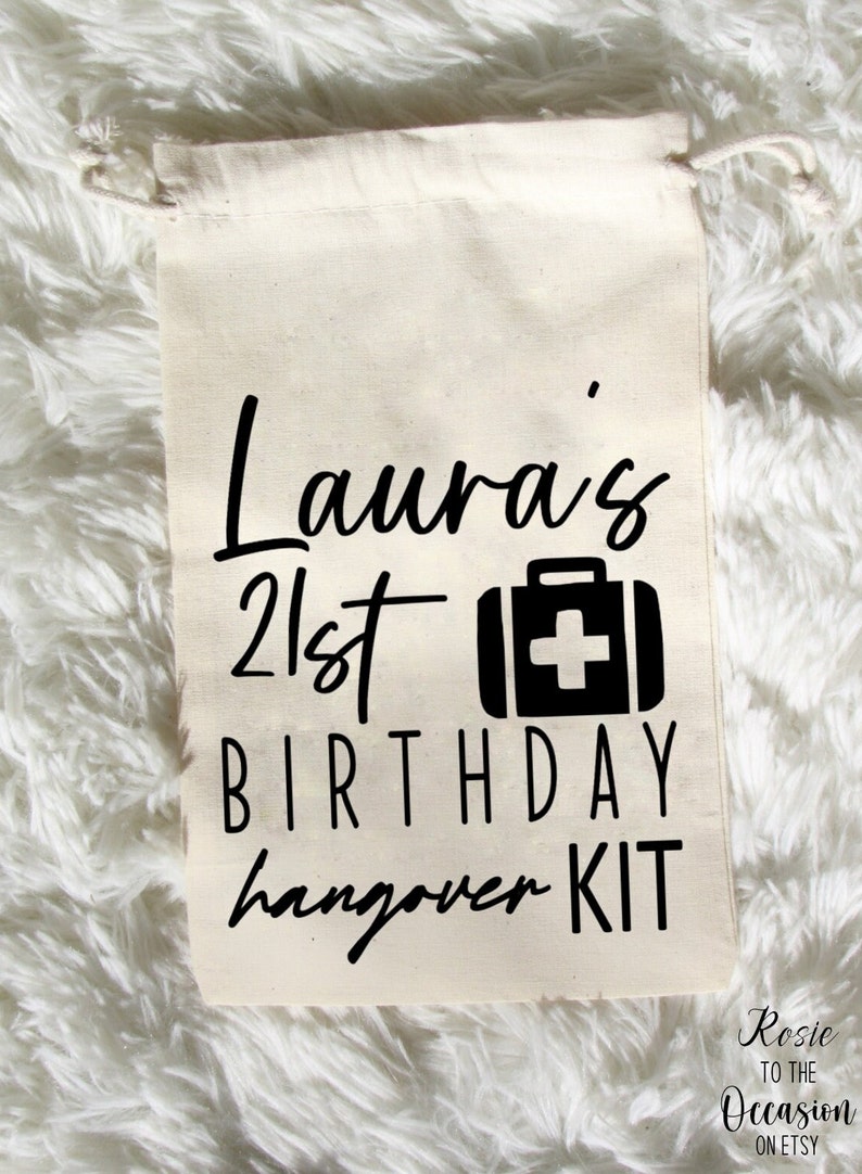 Op de afbeelding: Een witte trekkoordtas met zwarte tekst die "Laura's 21st Birthday Hangover Kit" luidt en een zwart EHBO-doosicoon.