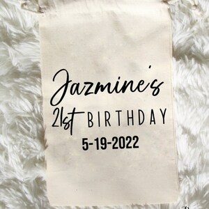 May include: A white cotton drawstring bag with black text that reads "Jazmine's 21st Birthday 5-19-2022". The bag is on a white fluffy surface.