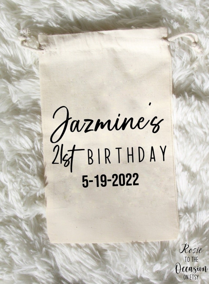 May include: A white cotton drawstring bag with black text that reads "Jazmine's 21st Birthday 5-19-2022". The bag is on a white fluffy surface.