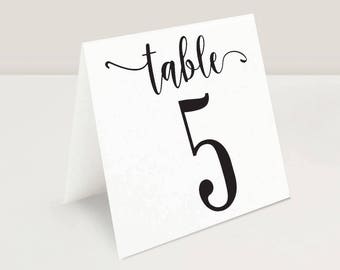 Könnte beinhalten: Eine weiße Tischnummerkarte mit dem Wort "table" in eleganter Schrift über der Zahl "5". Die Zahl ist in einer fetten, schwarzen Schriftart gehalten. Die Karte ist gefaltet und steht von selbst.