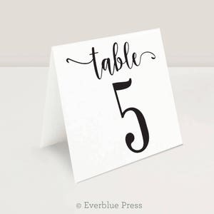May include: A white table number card with the word "table" in elegant script above the number "5". The number is in a bold, black font. The card is folded and stands on its own.