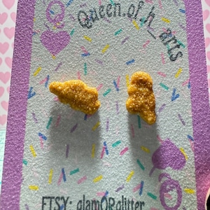 May include: A pair of novelty earrings shaped like chicken nuggets. The earrings are on a glittery, rectangular card with the text "Queen of Hearts" and "ETSY: glamORglitter". The card has a purple border and a background with hearts.