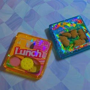 Könnte beinhalten: Zwei quadratische Kunstwerke aus Harz mit Miniatur-Lebensmitteln. Das orangefarbene Stück ist mit Lunch Ham & American Cracker Stackers beschriftet. Das blaue Stück trägt die Aufschrift Kid Cuisine Dino Nugget und 7 oz. Beide haben holografische Akzente.