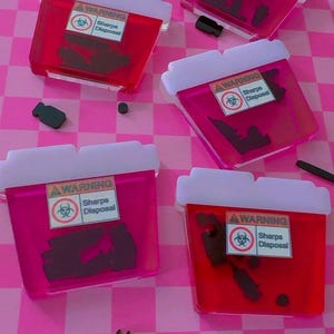 May include: Five translucent pink and red acrylic sharps disposal containers featuring white lids and a warning label that reads WARNING Sharps Disposal with a biohazard symbol. Each container holds various black acrylic shapes of medical tools.