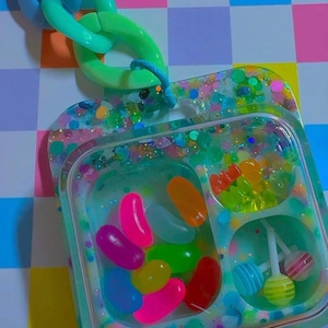 May include: A colorful resin tray with a variety of candies, including jelly beans and lollipops. The tray is filled with glitter and has a chain attached. The background is a checkered pattern of pink, blue, yellow, and orange squares.