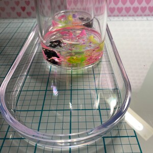 Puede incluir: Un recipiente de pl&aacute;stico transparente lleno de purpurina de colores, peque&ntilde;os amuletos negros y rosas y remolinos verdes y amarillos. El recipiente est&aacute; sentado en una bandeja de pl&aacute;stico transparente con un patr&oacute;n de cuadr&iacute;cula.
