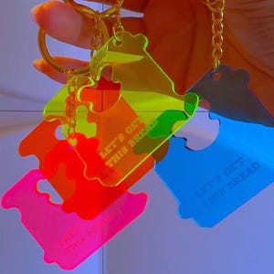 Puede incluir: Un conjunto de llaveros de acrílico translúcido en colores neón amarillo, naranja, rosa y azul, con forma de clip para pan. Cada llavero incluye una anilla y cadena de metal dorado, con el texto LET'S GET THIS BREAD grabado en la superficie.