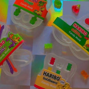 Könnte beinhalten: Draufsicht auf transparente Süßigkeitenformen mit bunten Gummibonbons und Haribo-Marken. Die Formen sind wie Frösche, Kirschen und Gummibärchen geformt. Der Hintergrund hat einen holografischen, irisierenden Effekt.