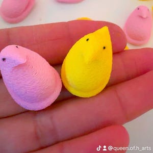 Op de afbeelding: Een hand die twee pastelkleurige marshmallow-snoepjes vasthoudt in de vorm van kuikens. Eén kuiken is geel en de andere is roze. De snoepjes zijn versierd met zwarte ogen.
