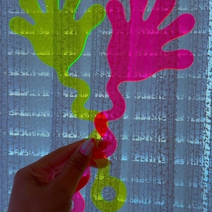 May include: Two translucent acrylic hand-shaped clappers, one in neon yellow and one in neon pink. Each clapper features a long, wavy handle ending in a circular loop. The vibrant, semi-transparent material allows light to pass through the shapes.