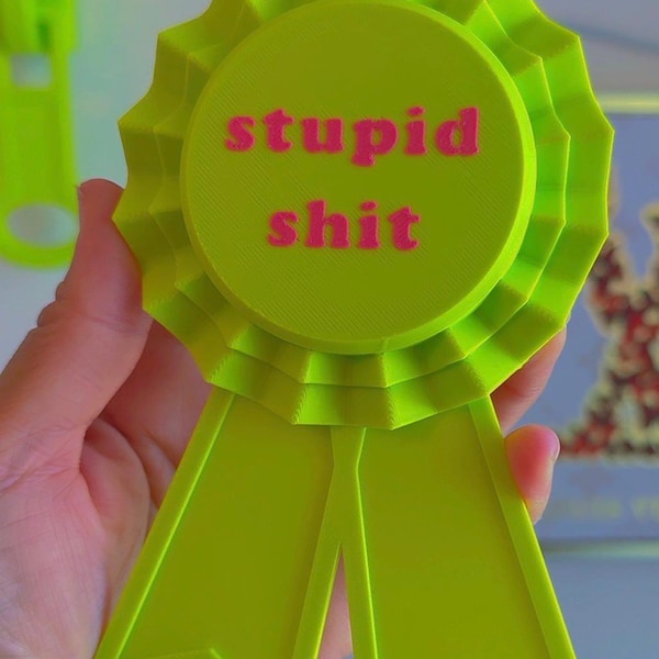 May include: A neon green award ribbon with the words "stupid shit" in pink. The ribbon has a circular center with a ruffled edge and two downward-pointing streamers. The text is centered on the circular portion of the ribbon.