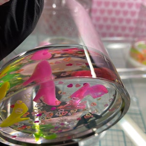 Puede incluir: Un tarro de vidrio transparente lleno de agua y purpurina de colores. La purpurina incluye estrellas y corazones rosas, amarillos, verdes y blancos. Tambi&eacute;n hay dos figuritas rosas dentro del tarro.